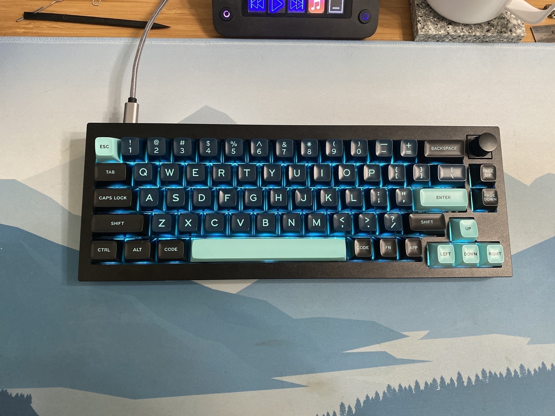 A Keychron Q2 keyboard with a turquoise backlight and teal/turquoise keycaps. Sitting on a blue lake/mountain scene deskpad.